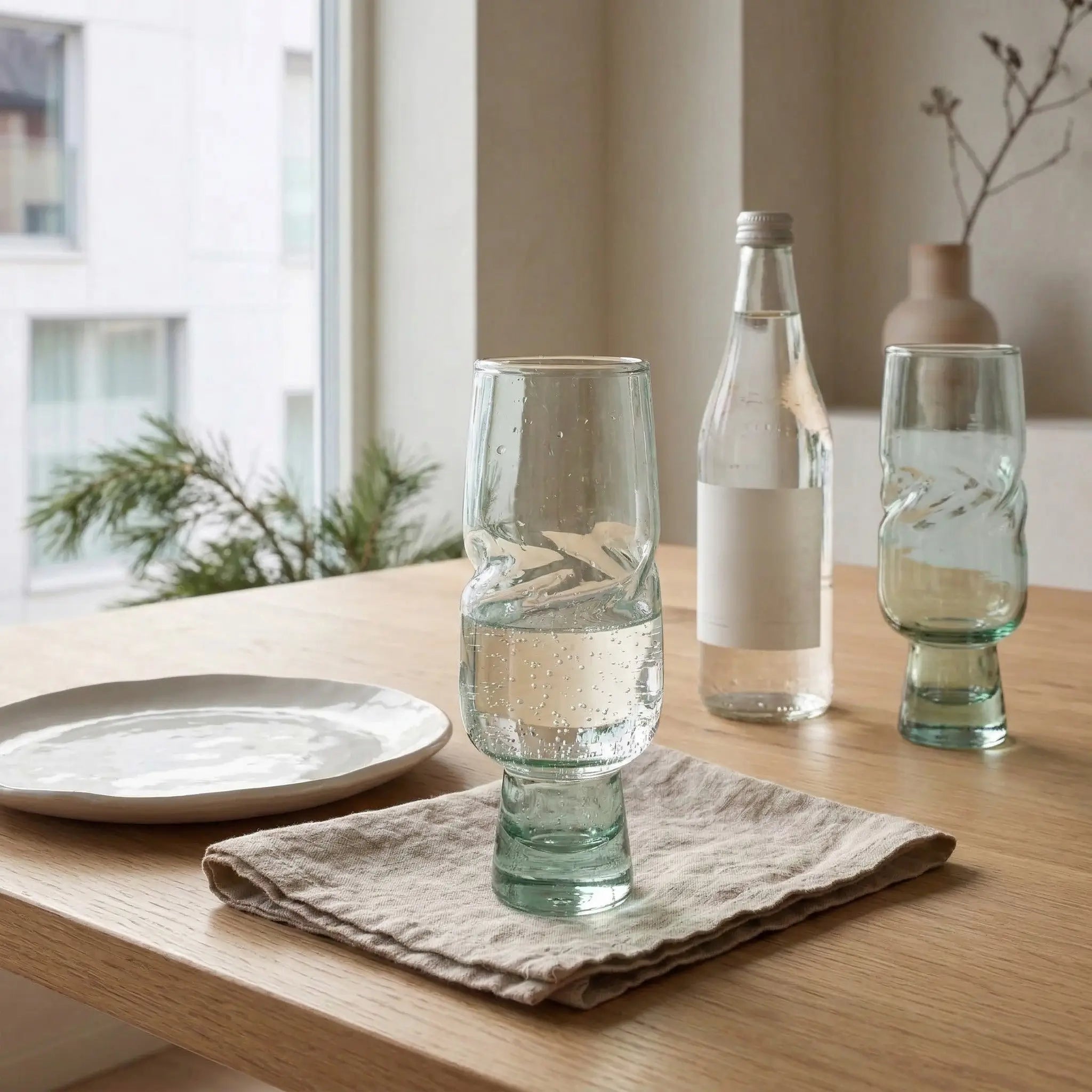 Clear VISLA glass with water on a wooden table with a bottle and another glass in the background.