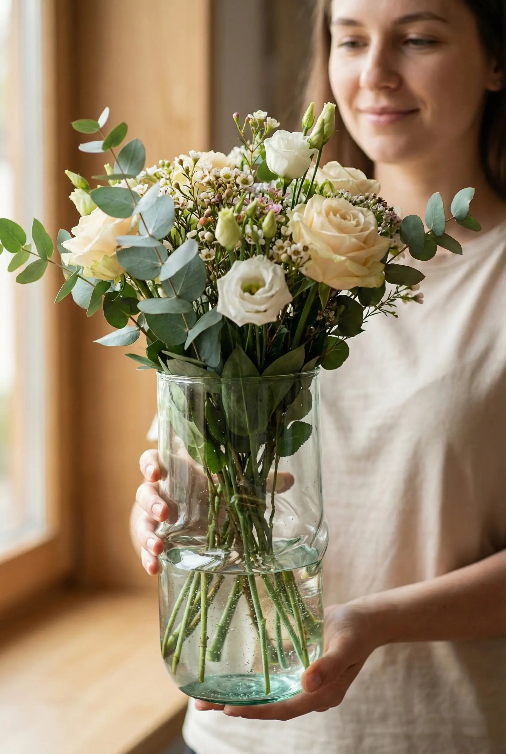Woman holding a bouquet of flowers in a clear vase indoors.
